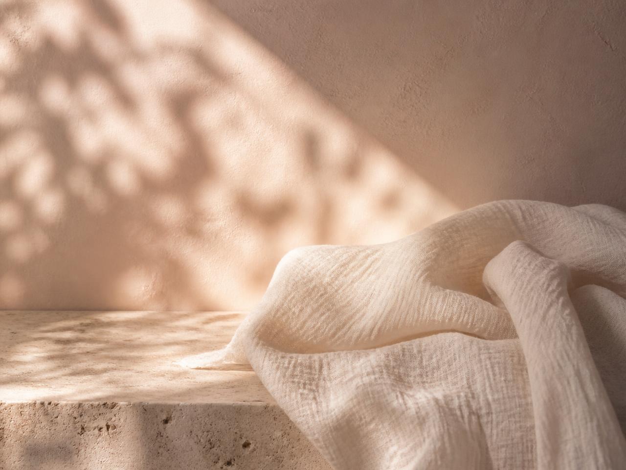Dappled sunlight across a warm plaster wall and a cream muslin cloth on travertine